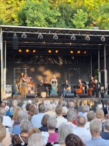 Bühne im Schlosspark Bisamberg mit Sängerin in grünem Kleid und Musikerinnen und Musikern, davor sitzendes Publikum