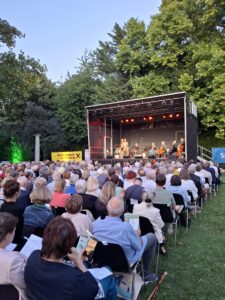 Bühne im Schlosspark Bisamberg mit Sängerin und Musikerinnen und Musikern, davor sitzendes Publikum