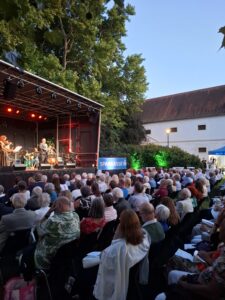 Bühne im Schlosspark Bisamberg mit Sängerin und Musikerinnen und Musikern, davor sitzendes Publikum