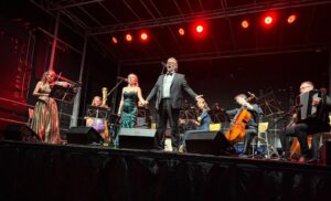 Sängerin in dunkelgrünem Kleid und Sänger in schwarzem Anzug händehaltend auf der Bühne. Dahinter Musikerinnen und Musiker